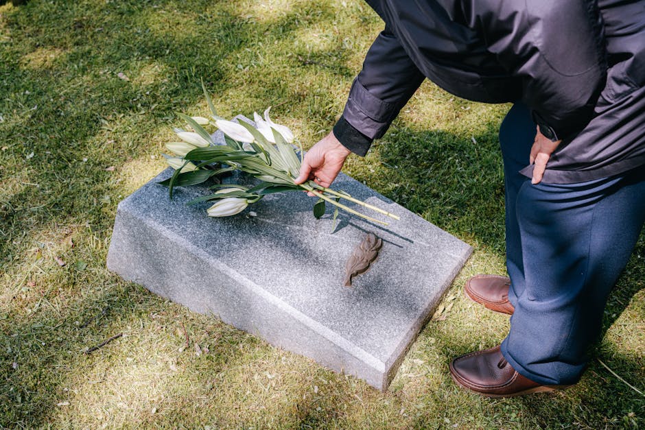 A person dressed in a dark jacket and blue trousers is placing a bouquet of white lilies with long green stems and leaves onto a grey granite grave marker at Kensal Green Cemetery. The bouquet is wrap