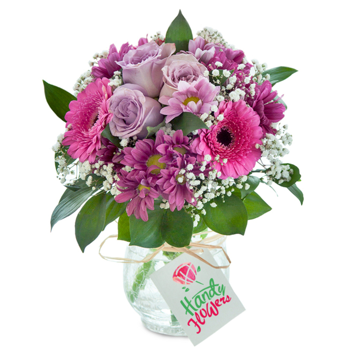 A vivacious medley of pink and purple florals captures the artistic pulse of Kensal Green in this thoughtfully arranged bouquet, spotlighted against a seamless white background. Central to the design, pale lavender roses gently unfurl their velvety petals, infusing the piece with classic romance and a soft, powdery fragrance evocative of morning air drifting over Kensal Rise. Large, vivid fuchsia Gerbera daisies radiate outward, their wide blossoms rimmed with striking dark centers, sparking visual energy akin to bustling Saturdays at the nearby farmer's market. Rich, deep magenta chrysanthemums - some with delicate yellow-green hearts - intermingle throughout, layering the arrangement with bold textures. Delicate sprays of pure white baby's breath hover above, adding refreshing lightness and a cloudlike charm that brightens the entire composition. The abundant blooms are anchored by fresh, glossy leaves, their deep green hues providing a soothing base. All elements are harmoniously nestled in a rounded clear glass vase featuring a charmingly crackled surface, reflecting the day's light in playful fragments. The finishing touch is a rustic raffia bow neatly tied at the neck, holding a crisp white gift tag with a hand-drawn floral motif - a subtle signature of Kensal Green's dedication to both craft and local community. Perfect for birthdays, heartfelt thank-yous, or spontaneous delights, this arrangement channels both the creative diversity and welcoming atmosphere of Kensal Green's neighbourhoods.
